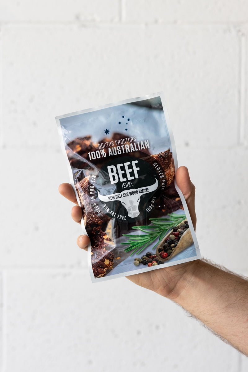 Hand holding large pack of wood smoked beef jerky up in the air in front of a white brick wall