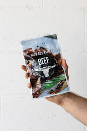 Hand holding large pack of wood smoked beef jerky up in the air in front of a white brick wall
